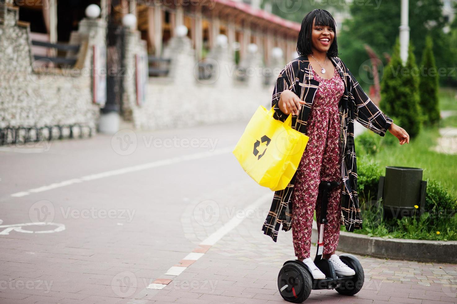 vacker afrikansk amerikansk kvinna som använder segway eller hoverboard. svart flicka med gul duk eco väskor återvinning symbol. foto