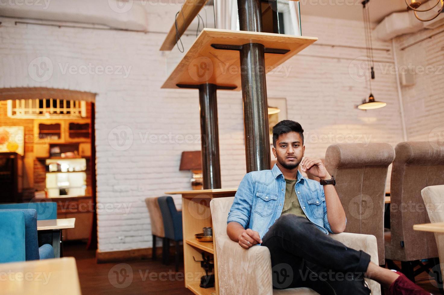 porträtt av stilig framgångsrik skäggig sydasiatisk, ung indisk frilansare i blå jeansskjorta som sitter på café och vilar. foto