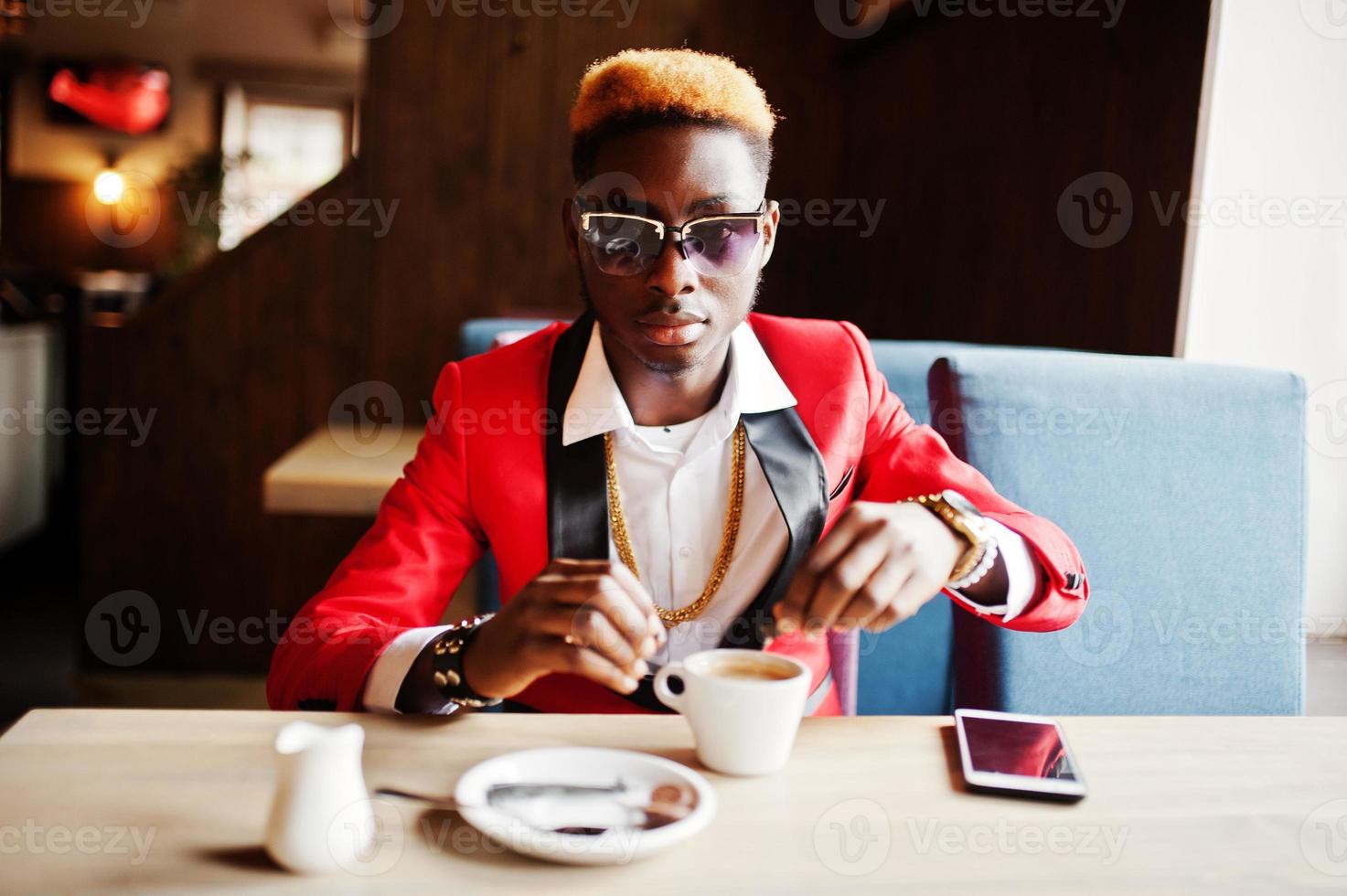 mode afrikansk amerikansk man modell i röd kostym, med höjdpunkter hår och solglasögon sitter på café med kaffe. foto