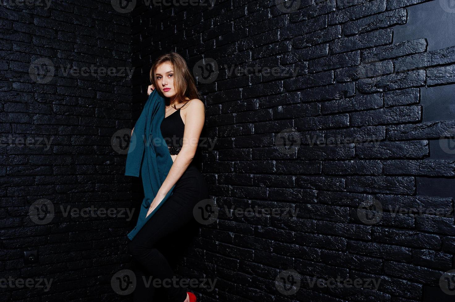 stilig brunett flicka bära på svart, poserar på studio mot mörk tegelvägg. studio modell porträtt. foto