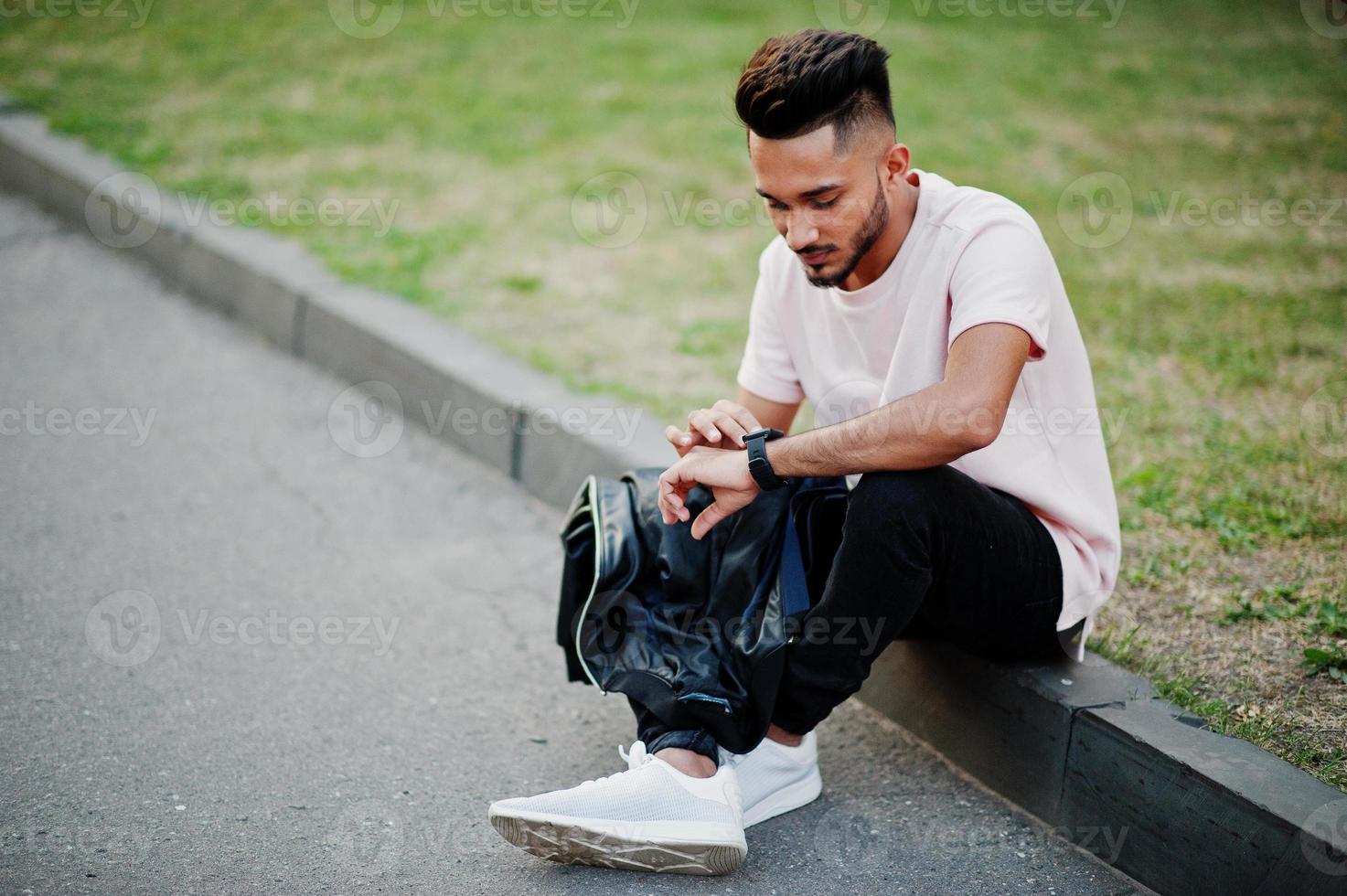 snygg indisk skäggman på rosa t-shirt. indisk modell sitter vid gröna trädgården i staden och tittar på sina moderna klockor. foto