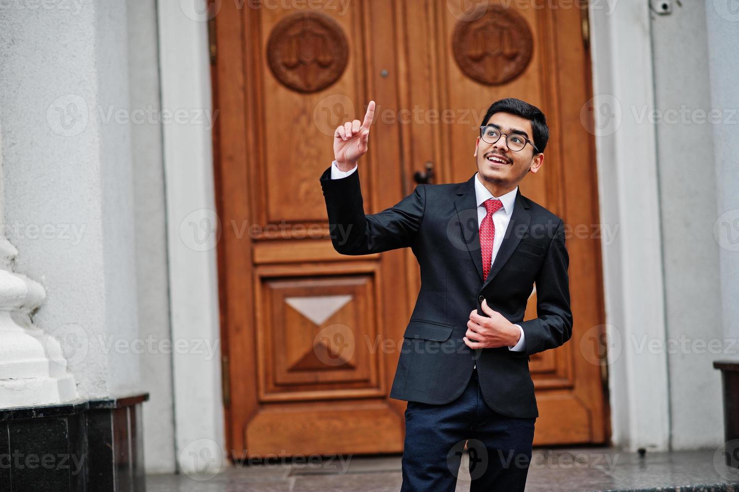 indisk ung man på glasögon, bära på svart kostym med röd slips poserade utomhus mot dörren till byggnaden visa finger upp, han har idé. foto