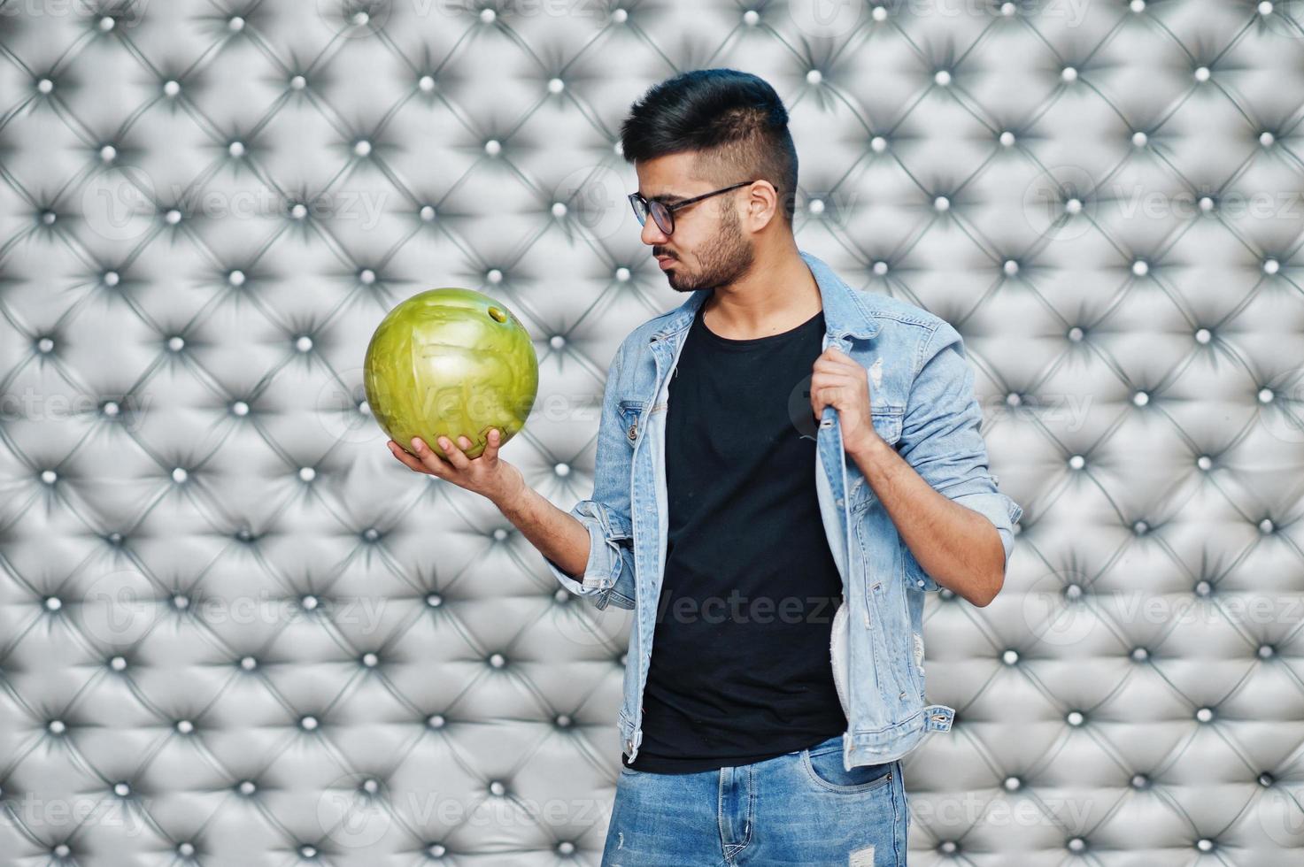 snygg asiatisk man i jeansjacka och glasögon med bowlingklot till hands mot silverväggbakgrund. foto