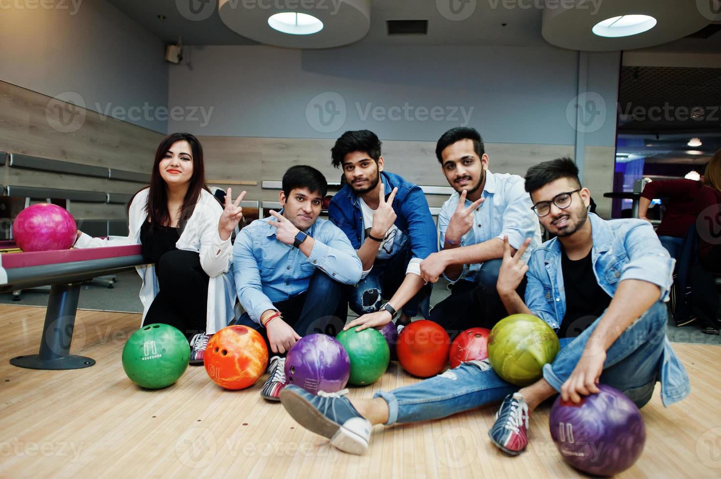 grupp av fem sydasiatiska folk som har vila och roligt på bowlingklubben. håller bowlingklot, sitter på gränden och visar två fingrar tecken. foto