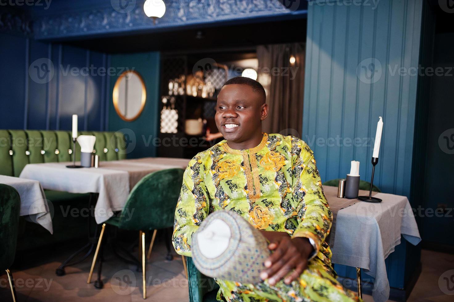 vänlig afro man i traditionella gula kläder sitter på restaurang och håller mössa till hands. foto