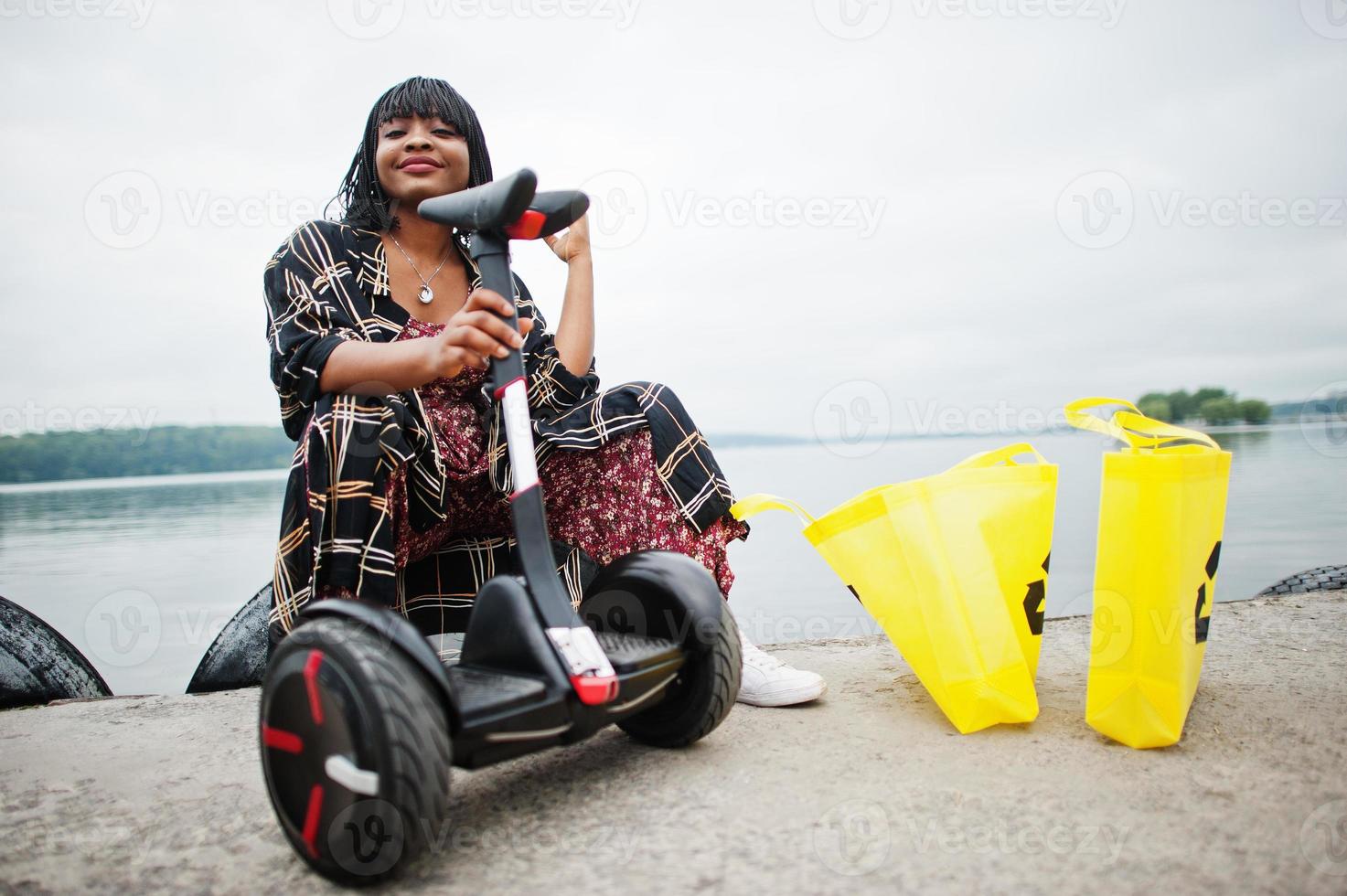 vacker afrikansk amerikansk kvinna sitter nära segway eller hoverboard. svart flicka med gul duk eco väskor återvinning symbol. foto