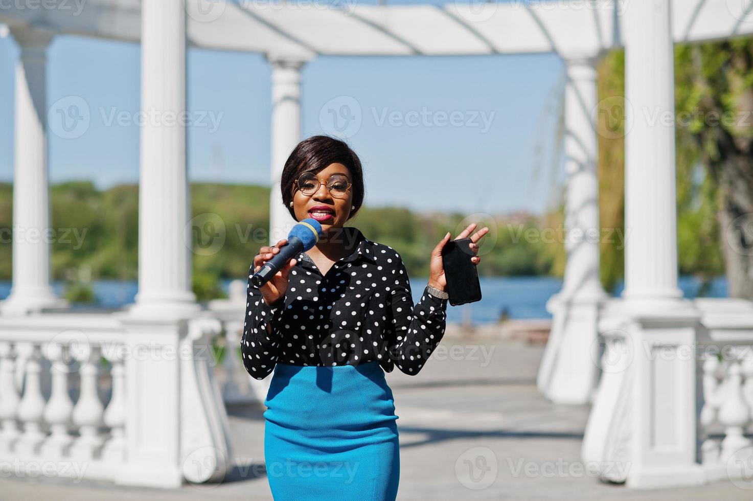 snygg afrikansk amerikansk modell tjej tv-presentatör med mikrofon i glasögon, blå kjol och svart blus poserade utomhus mot vit stenbåge med mobiltelefon. foto