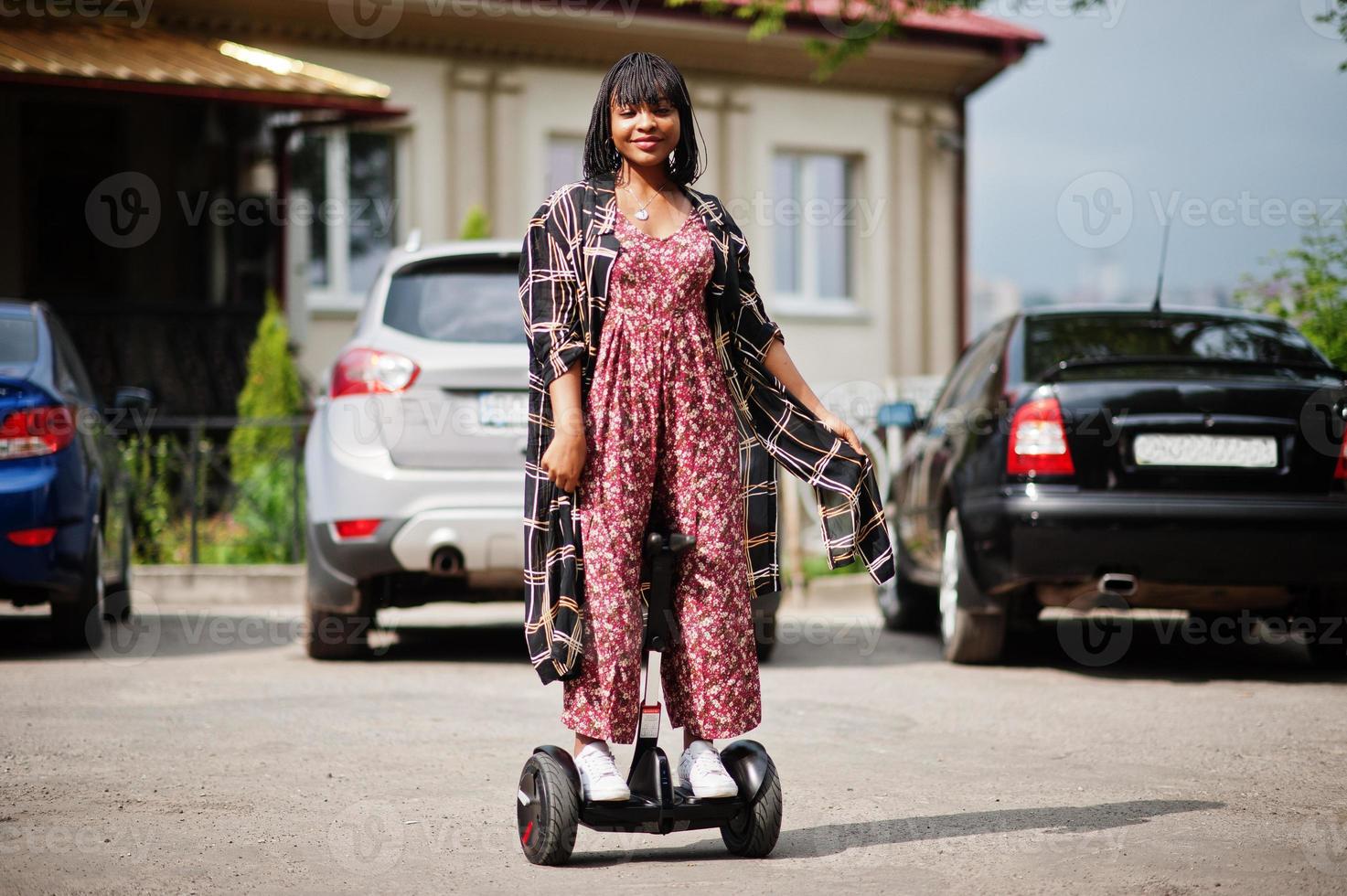 vacker afrikansk amerikansk kvinna som använder segway eller hoverboard. svart flicka på dubbla hjul självbalanserande elektrisk skoter mot bilparkering. foto