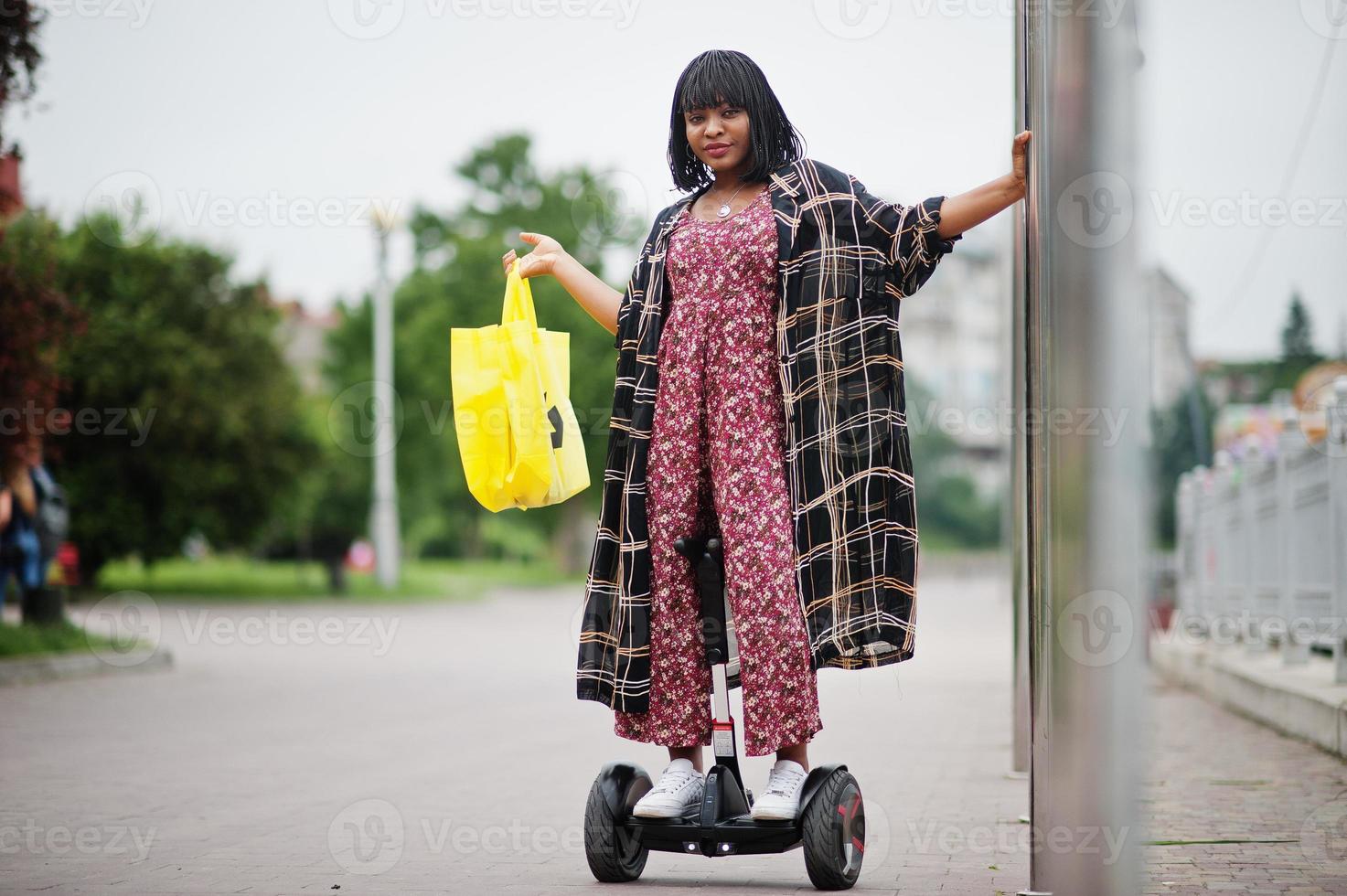 vacker afrikansk amerikansk kvinna som använder segway eller hoverboard. svart flicka med gul duk eco väskor återvinning symbol. foto