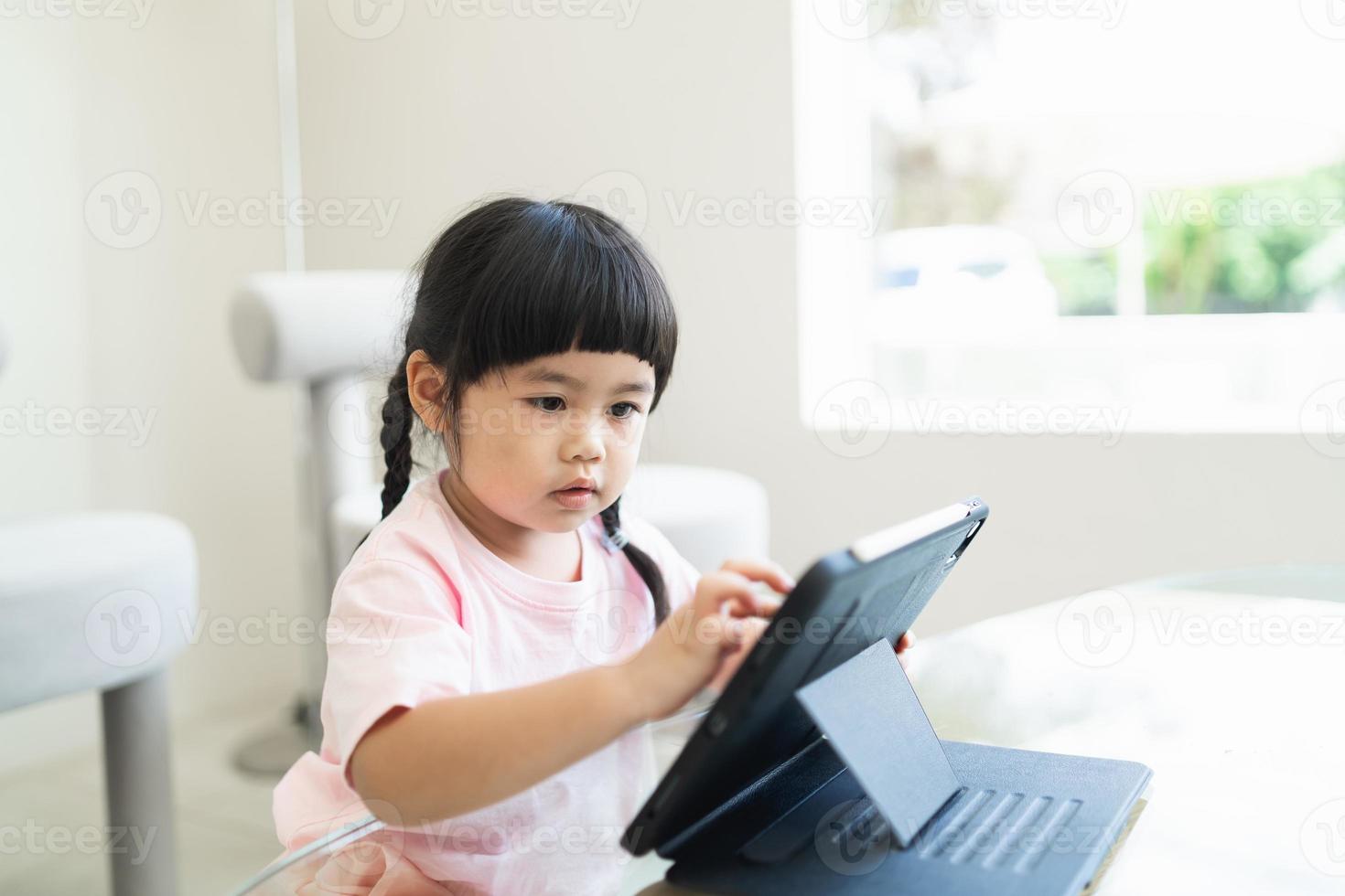 asiatisk tjej tittar och trycker på surfplattans skärm uppmärksamt. överstimulerade barn koncept. för mycket skärmtid. asiatisk tjej som tittar på videor medan tv:n arbetar. foto