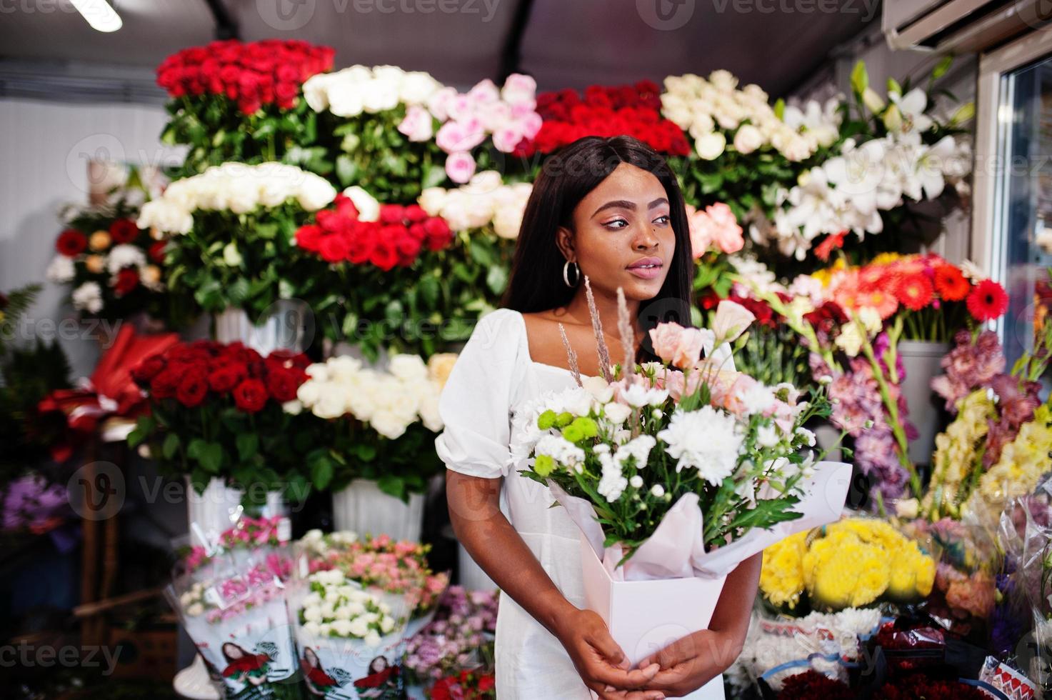 vacker afrikansk amerikansk flicka i öm vit klänning med bukettblommor i händerna står mot blommig bakgrund i blomsteraffär. svart kvinnlig florist. foto