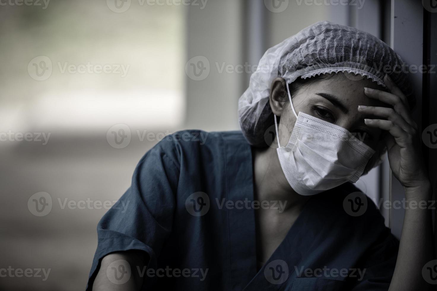 trött deprimerad kvinnlig asiatisk skrubbsköterska bär ansiktsmask blå uniform sitter på sjukhusgolvet, ung kvinna läkare stressad av hårt arbete foto