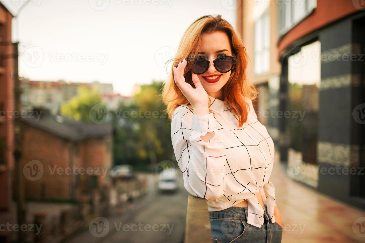 attraktiv rödhårig kvinna i solglasögon, bär på vit blus poserar på gatan mot modern byggnad. foto