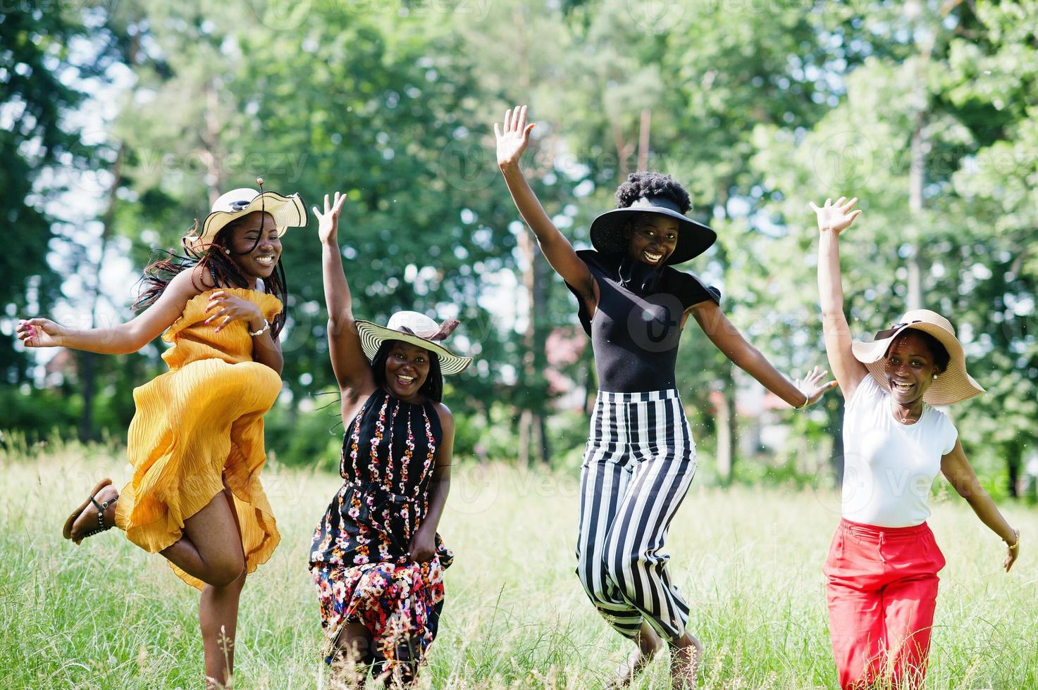 grupp av fyra underbara afroamerikanska kvinnor bär sommarhatt hoppar på grönt gräs i parken. foto