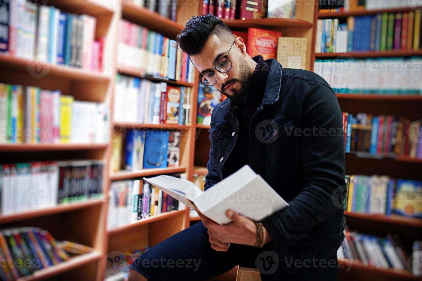 lång smart arabisk studentman, bär på svart jeansjacka och glasögon, på biblioteket med bok vid händerna. foto