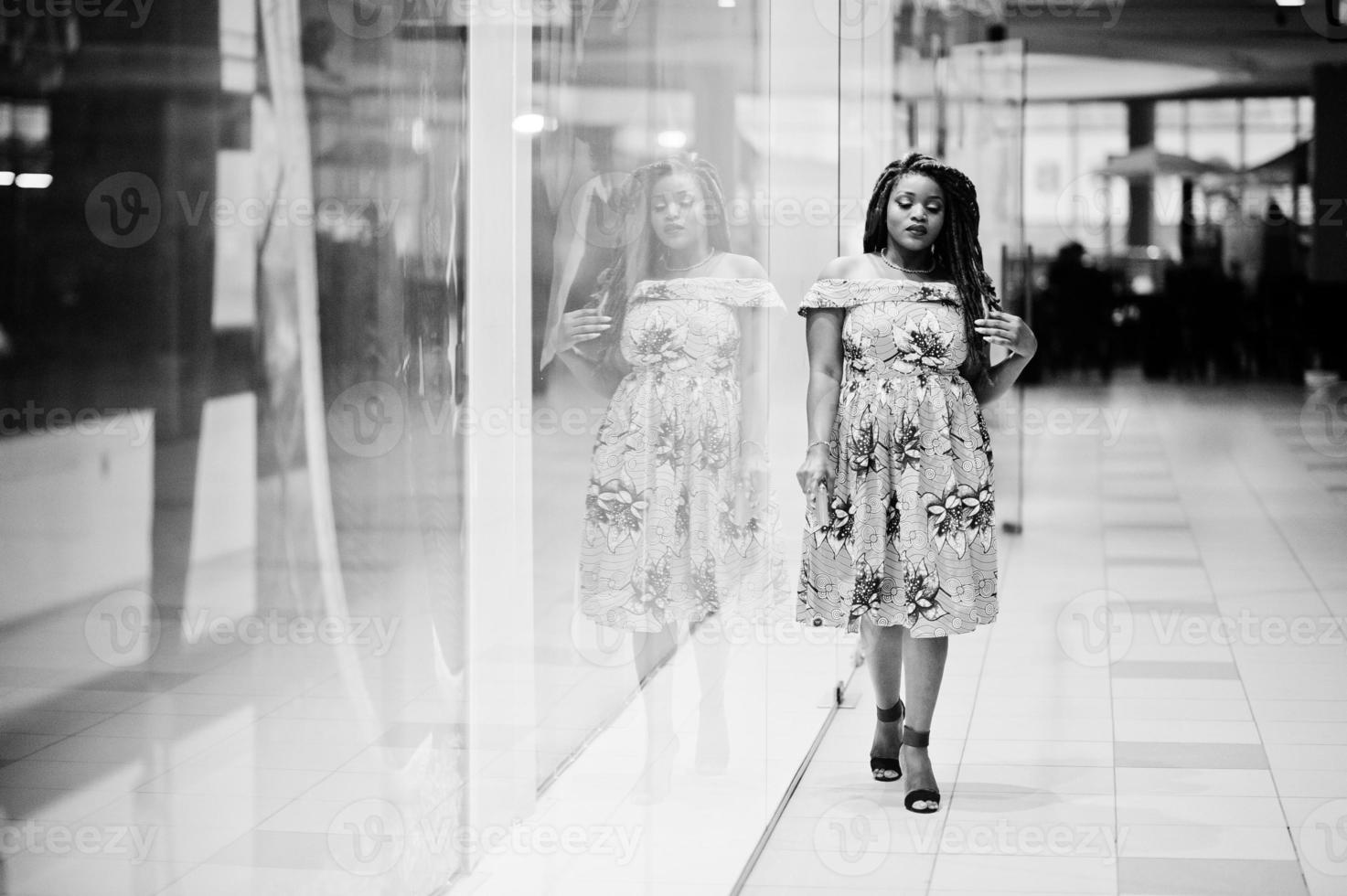 söt liten höjd afroamerikansk flicka med dreadlocks, bära på färgad gul klänning, poserade av butiken showcase på handelscentrum. svartvitt. foto
