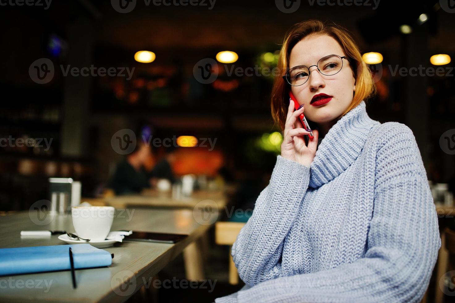 glad ung vacker rödhårig kvinna i glasögon som sitter på sin arbetsplats på café och pratar i mobiltelefon. foto