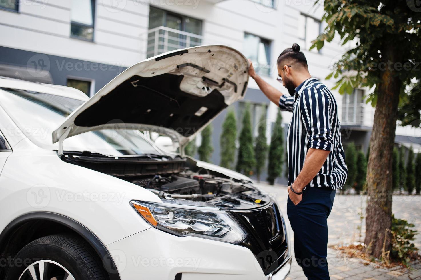 framgångsrik arab man bär i randig skjorta och solglasögon poserar nära sin vita suv bil, kontrollera motor med öppen huva. foto