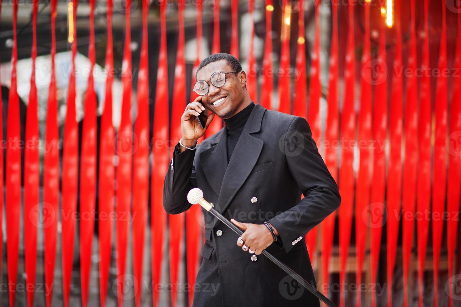 stilig afrikansk amerikansk herre i elegant svart jacka, med retro käpp som käppkolv eller tippande käpp. rik fashionabla afro man mot röd vägg talar i mobiltelefon. foto