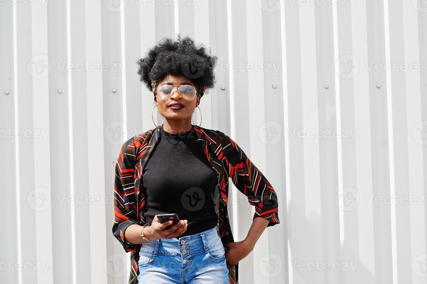 afrikansk kvinna med afro hår, i jeans shorts och glasögon poserade mot vit stålvägg med mobiltelefon i handen. foto