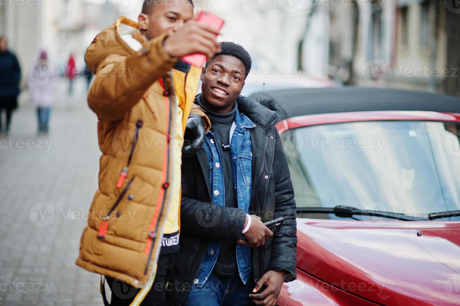 två afrikanska manliga vänner står nära röd amerikansk cabrioletbil och gör selfie på telefonen. foto