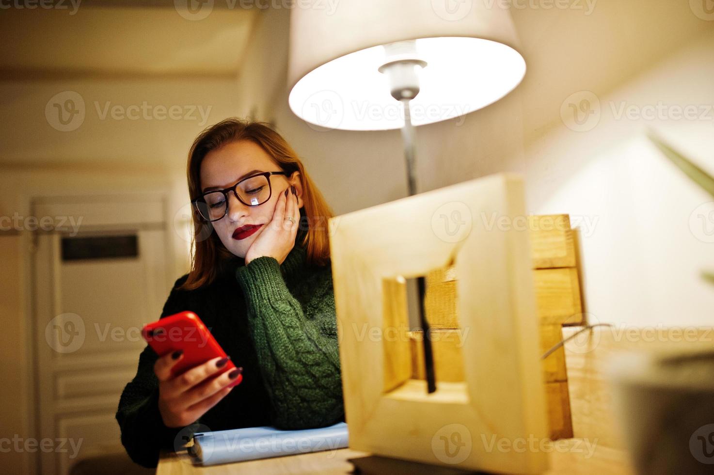 glad ung vacker rödhårig kvinna i glasögon, bär på grön varm ulltröja, sitter på café och använder sin mobiltelefon. foto