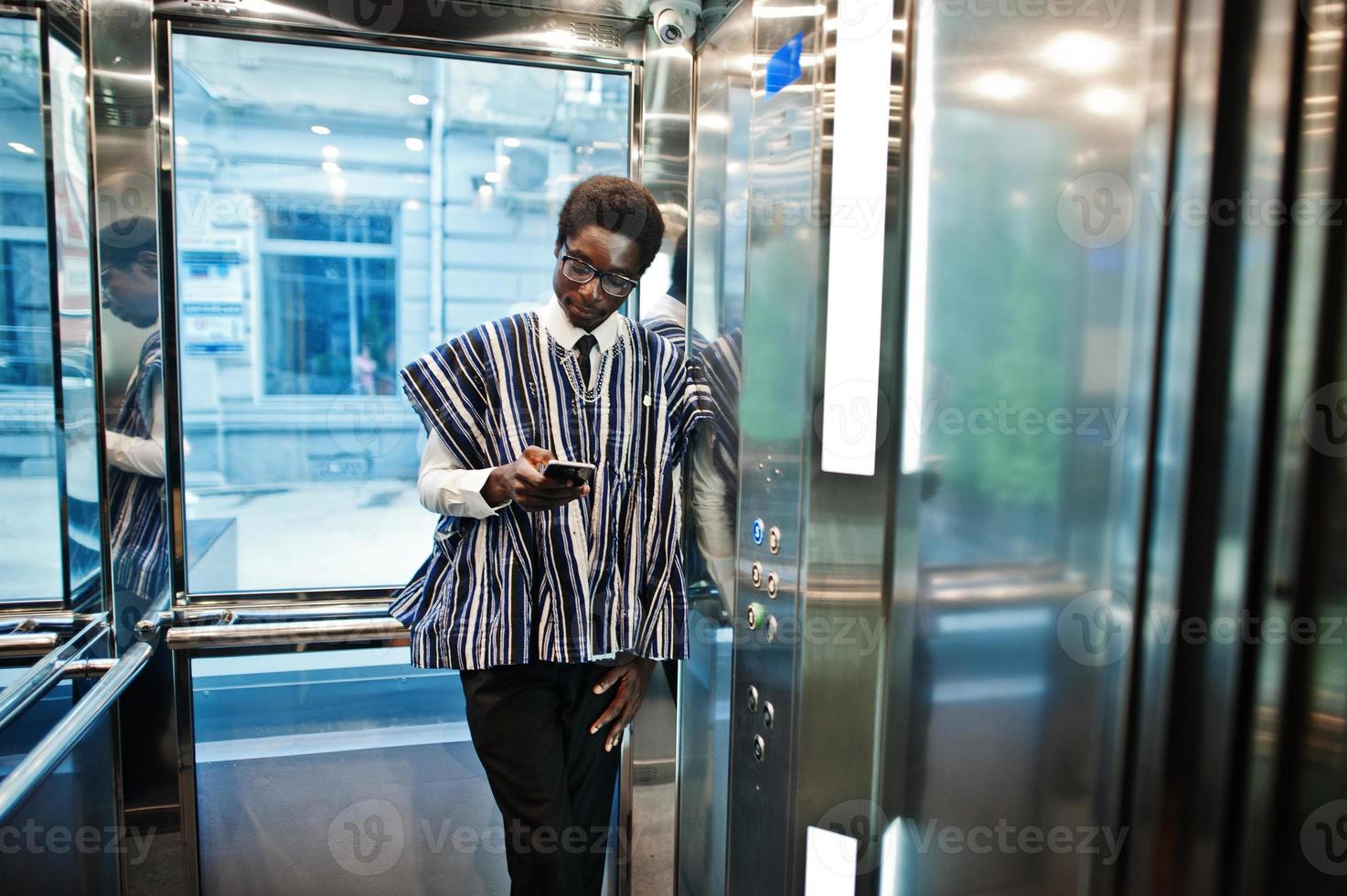 afrikansk man i traditionella kläder och glasögon med mobiltelefon vid hissen eller modern hiss. foto
