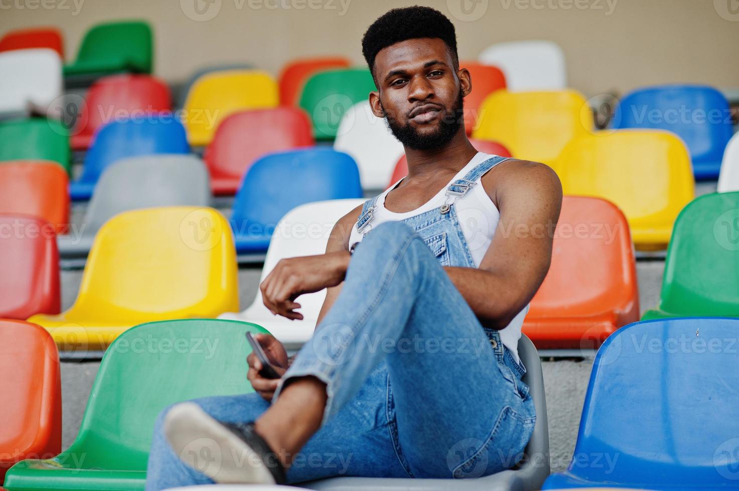 stilig afrikansk amerikansk man på jeansoveraller poserade på färgade stolar på stadion. fashionabla svart man porträtt. foto