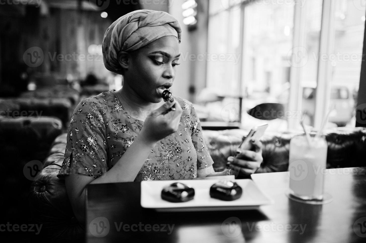 snygg afrikansk kvinna i röd skjorta och hatt poserade inomhuscafé, äter chokladdessertkakor och tittar på mobiltelefonen. foto