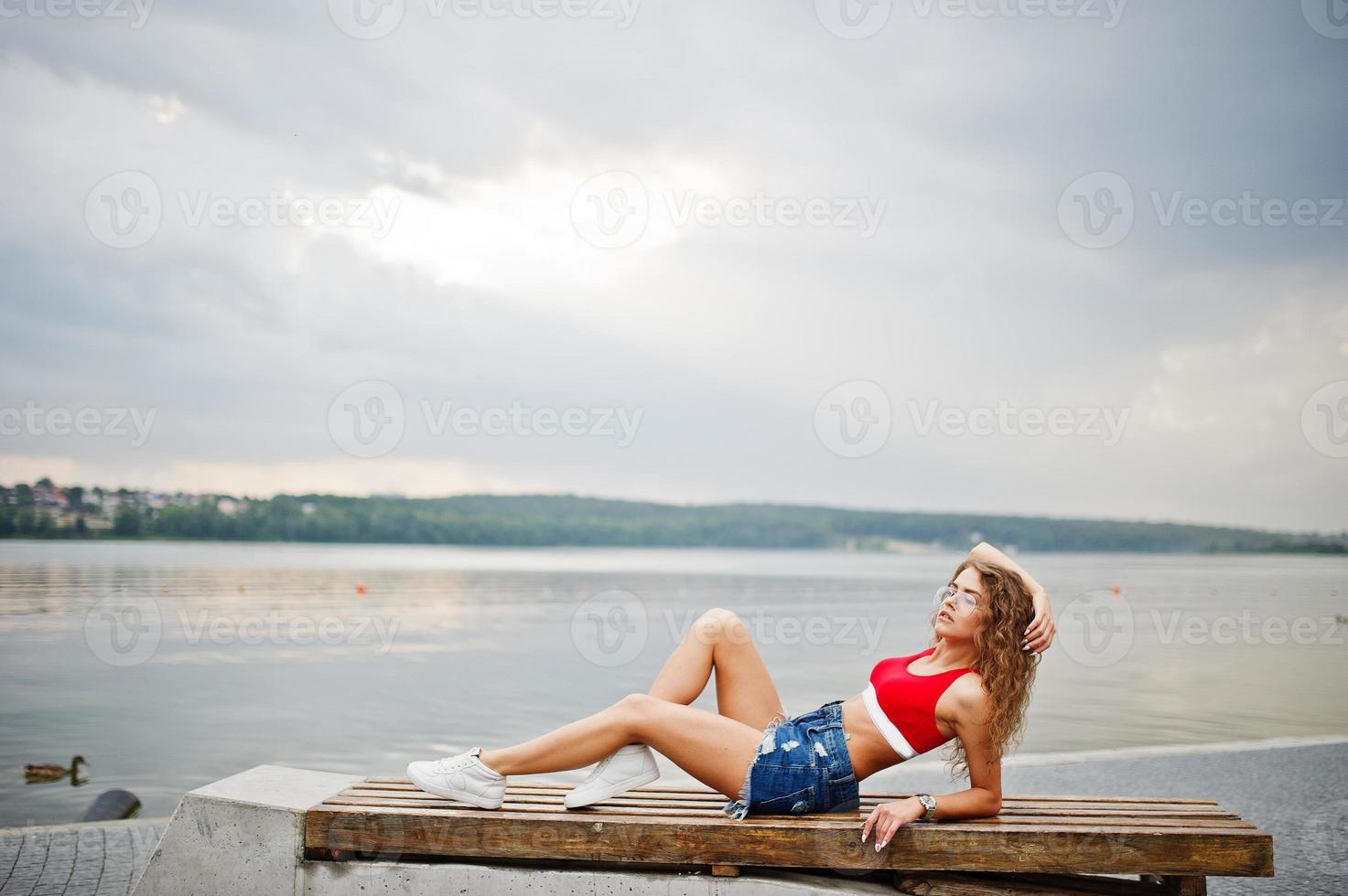 sexig lockig modell tjej i röd topp, jeans jeansshorts, glasögon och sneakers poserade på bänken mot sjön. foto