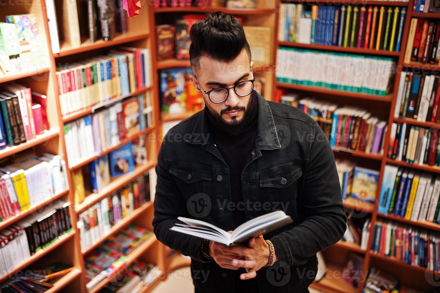 lång smart arabisk studentman, bär på svart jeansjacka och glasögon, på biblioteket med bok vid händerna. foto