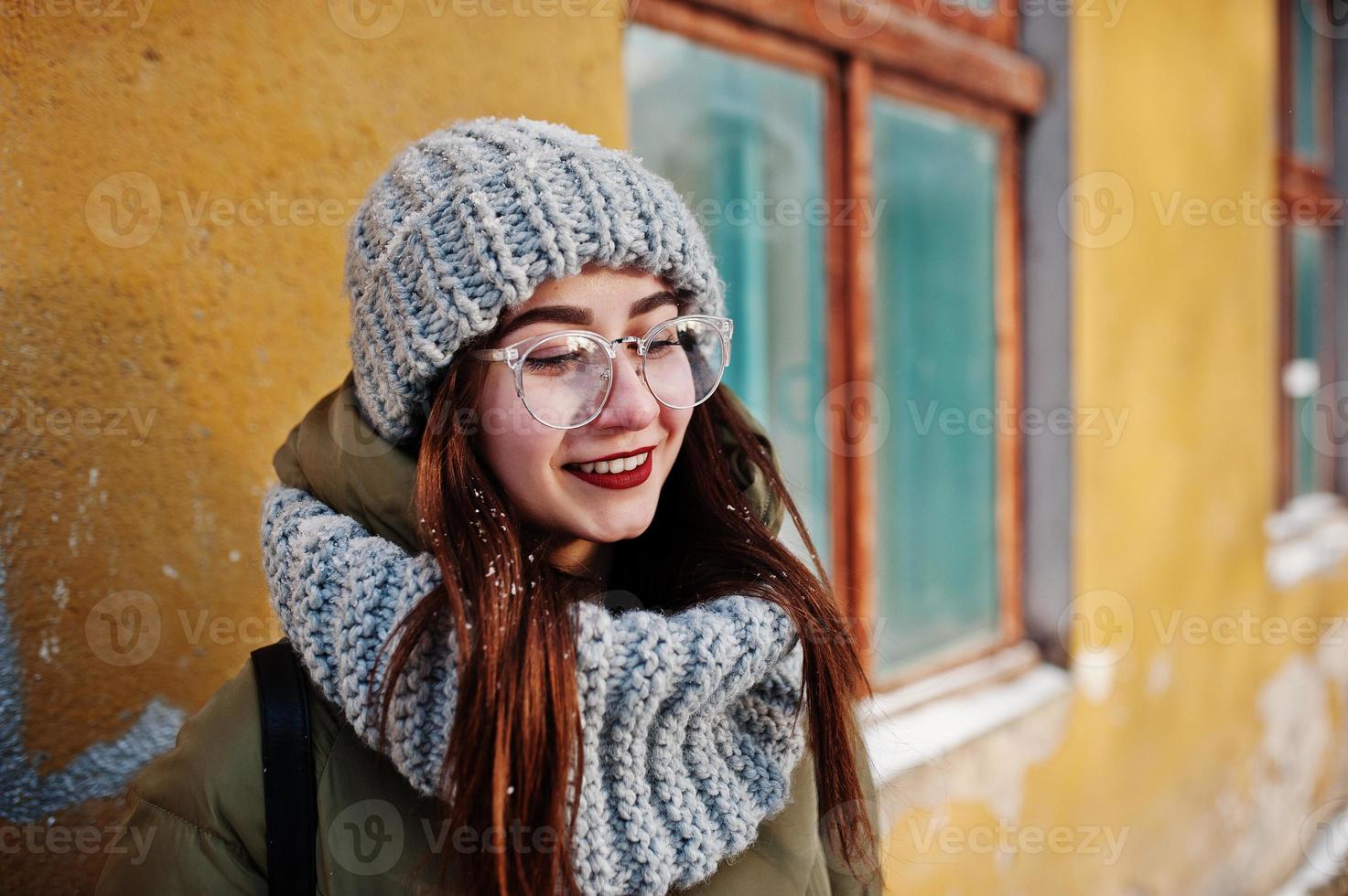porträtt av brunett flicka i grå halsduk och hatt, glasögon vid kallt väder med solsken mot orange vägg av gamla hus. foto