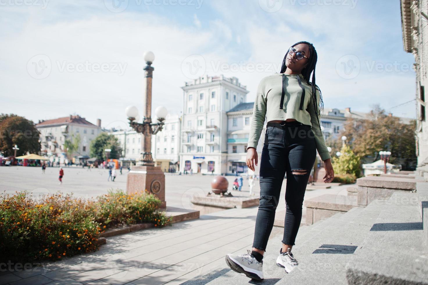 stadsporträtt av positiv ung mörkhyad kvinna som bär grön huvtröja och glasögon. foto