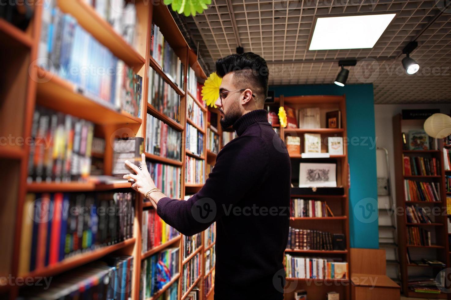 lång smart arabisk student man, bära på violett polotröja och glasögon, på biblioteket välja bok på hyllor. foto