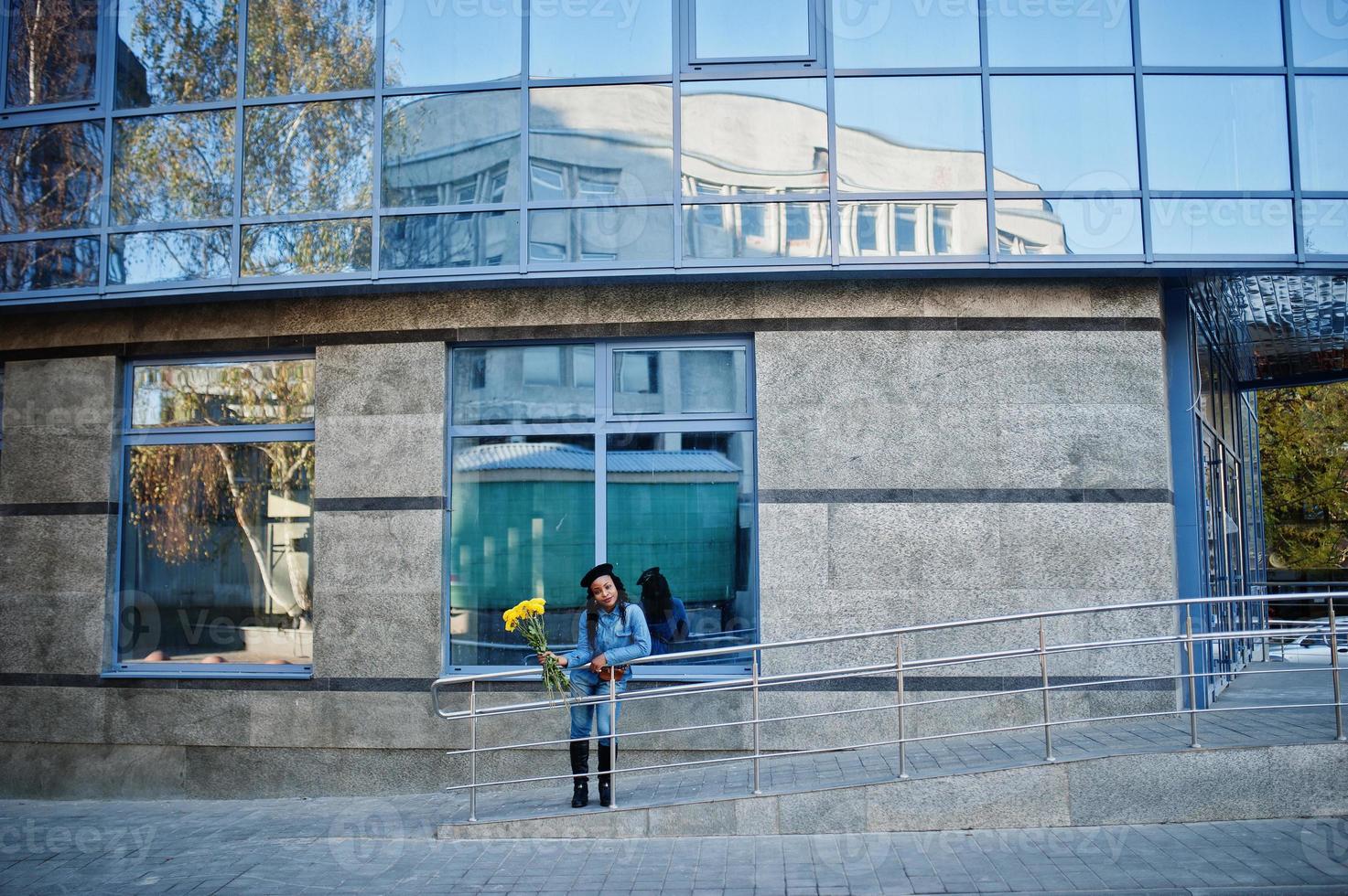 eleganta fashionabla afroamerikanska kvinnor i jeans och svart basker med gula blommor bukett poserade utomhus i solig dag mot blå modern byggnad. foto