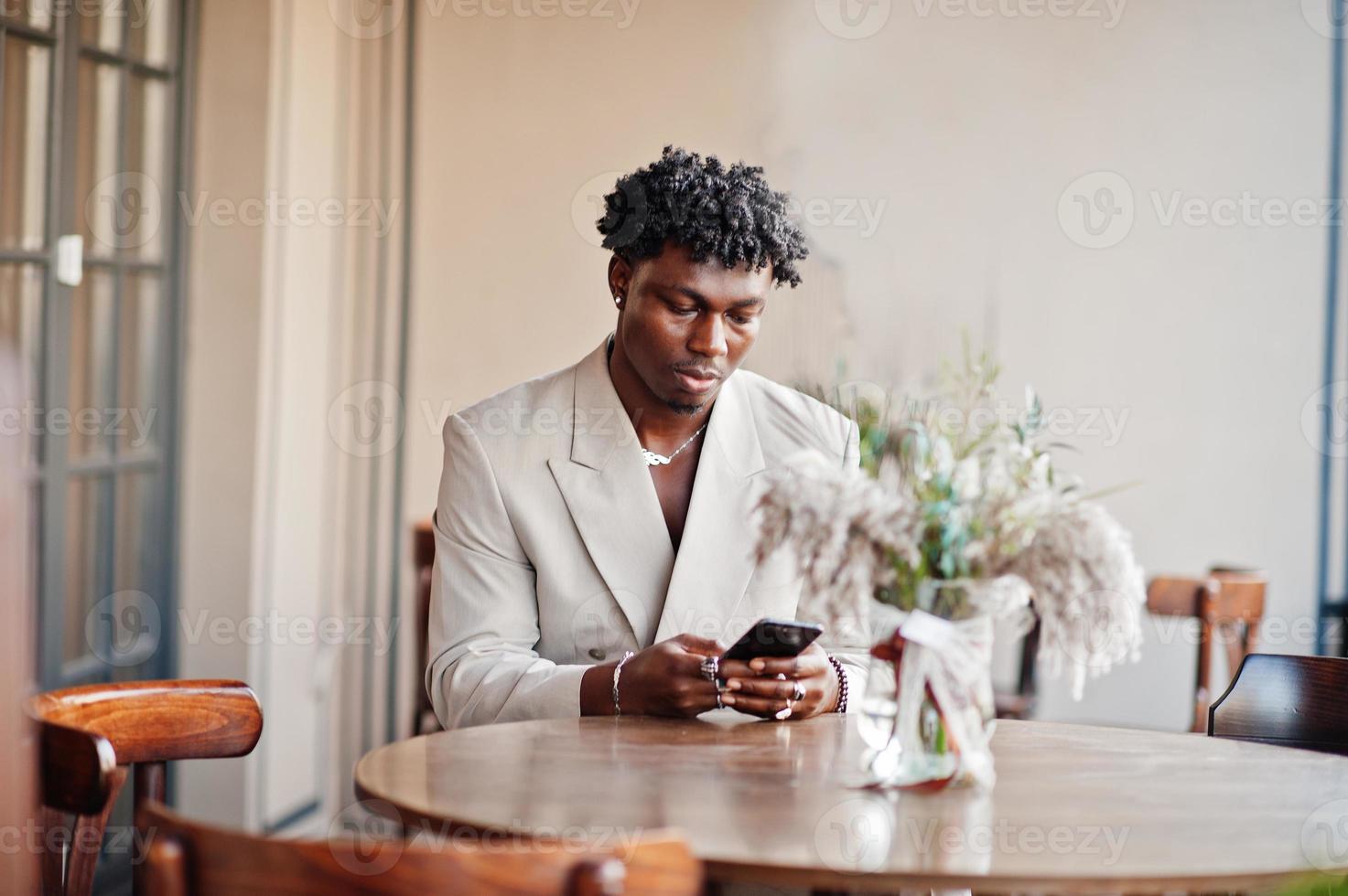 snygg afro man i beige gammal skolkostym sitter på café med mobiltelefon. fashionabla ung afrikansk man i ledig jacka på bar överkropp. foto