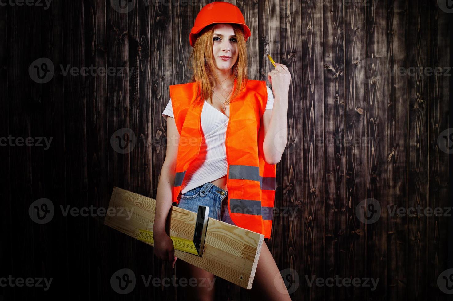 ingenjör kvinna i orange skydda hjälm och bygga jacka mot trä bakgrund håller styrelse och linjal. foto