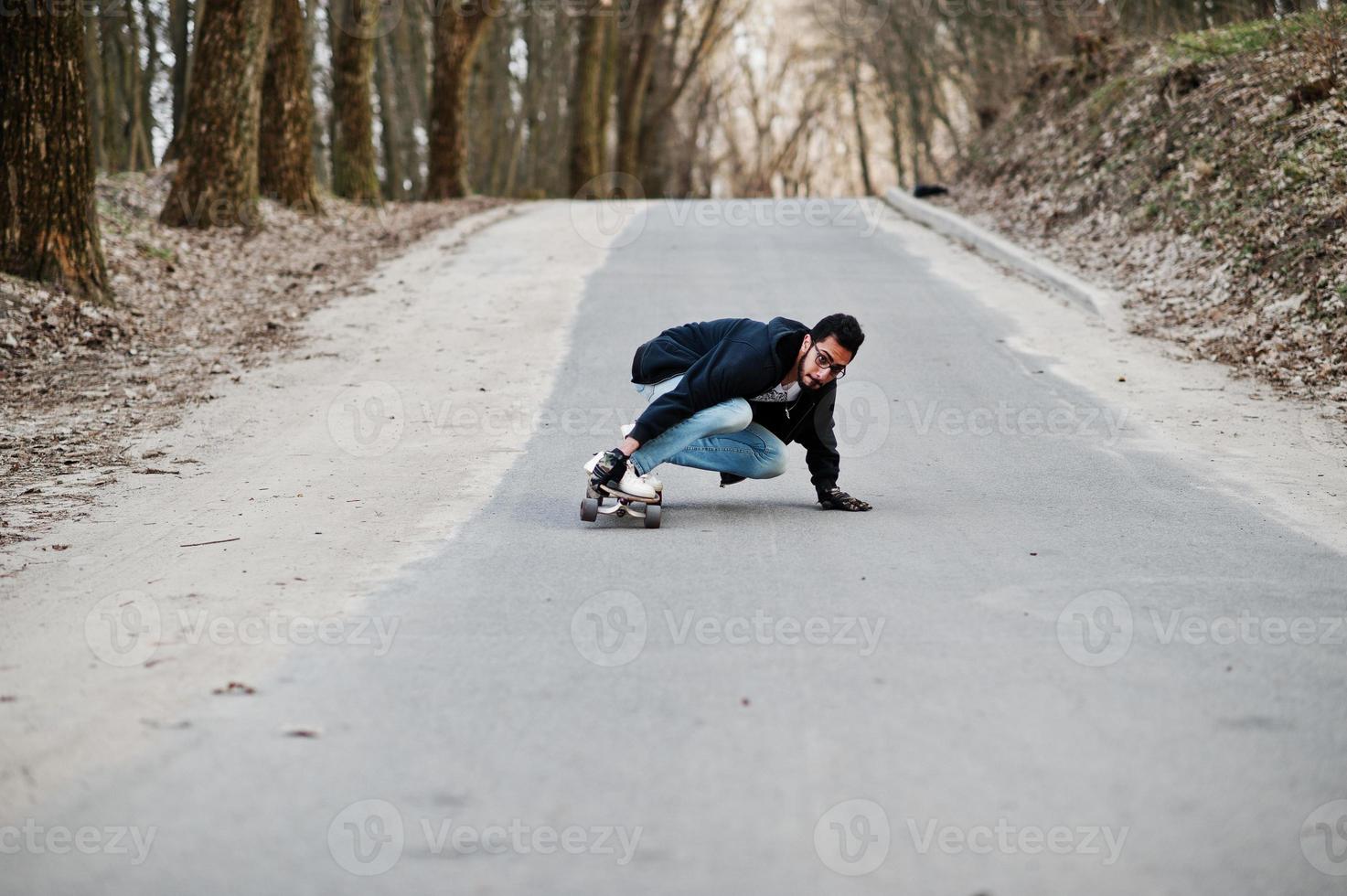 street style arabisk man i glasögon med longboard longboarding på vägen. foto