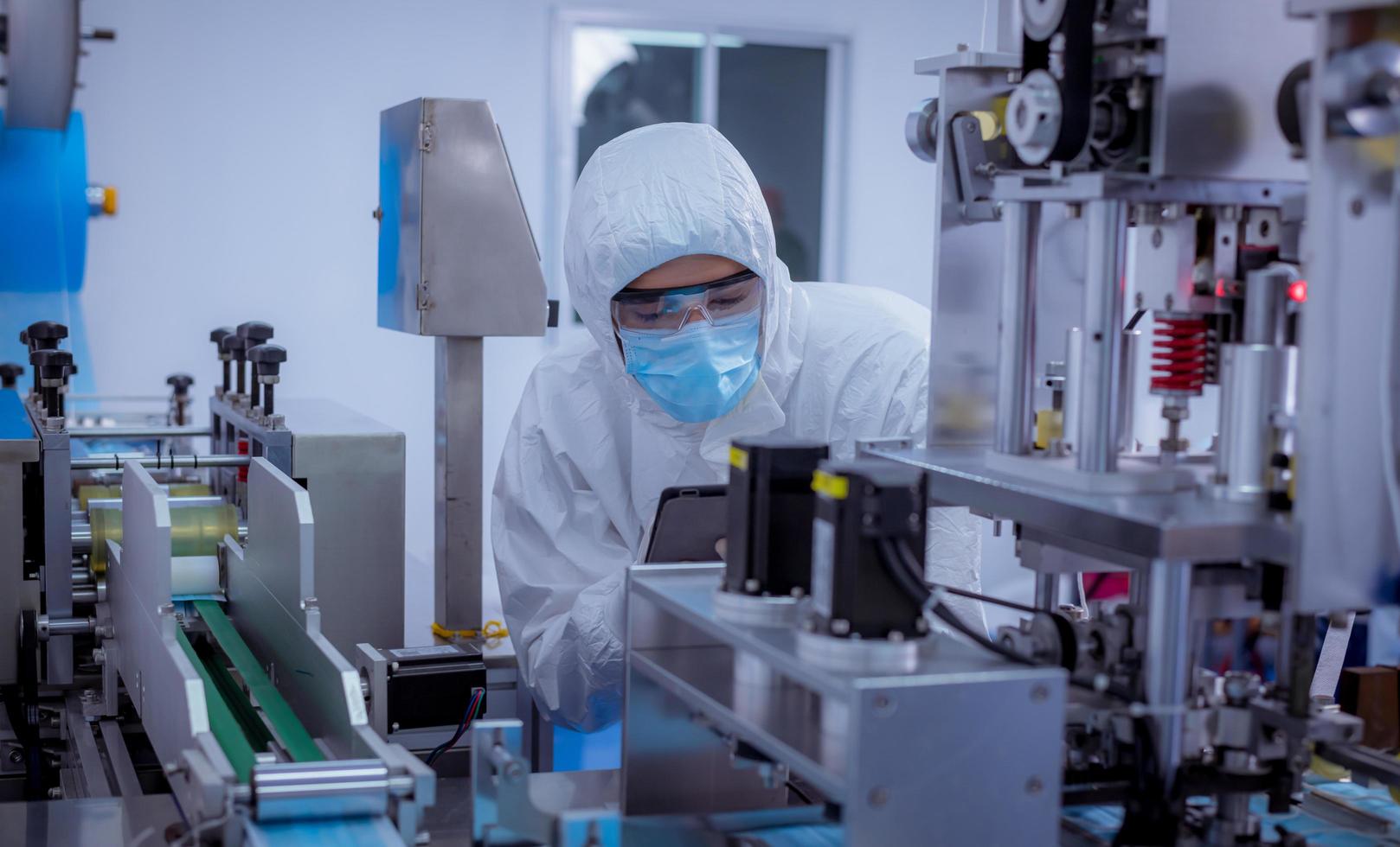 tekniker arbetare bär skyddssvit, handskar är inspektera maskinen med kontrollera ansiktsmask tillverkningsprocessen för att vara säker och mot virus foto