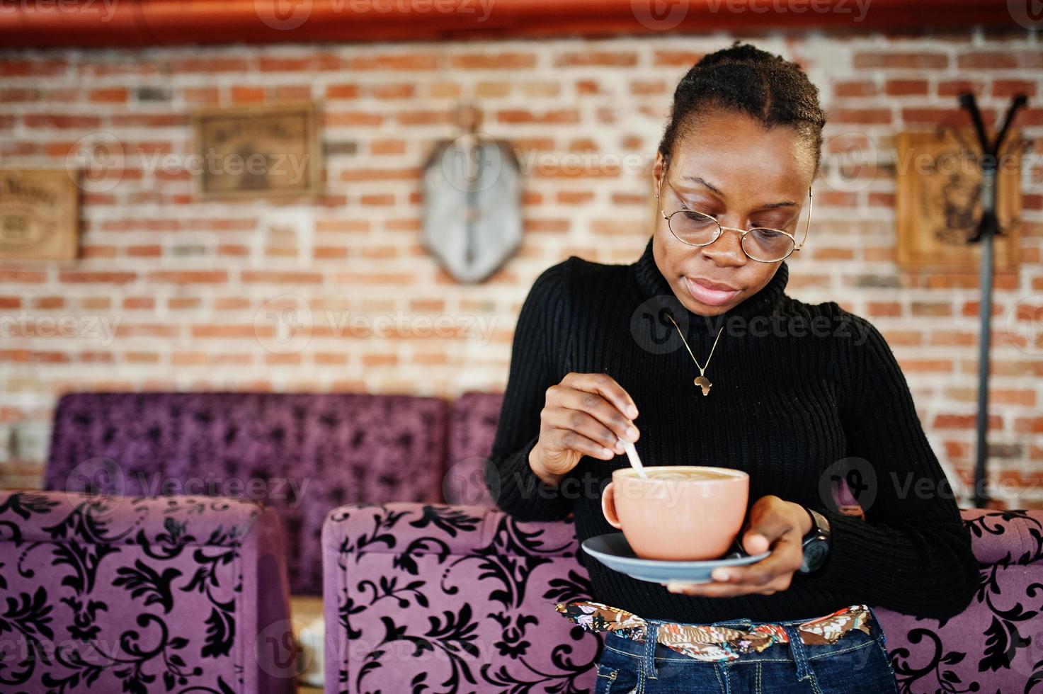 afrikansk kvinna i svart tröja och glasögon poserade på café med en kopp varm dryck. foto