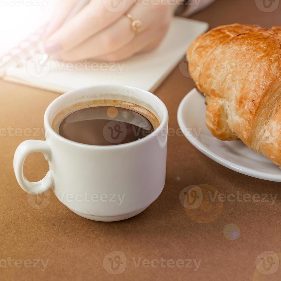 närbild av croissant och espresso. kvinna skriver på anteckningsboken. frilansare på jobbet foto