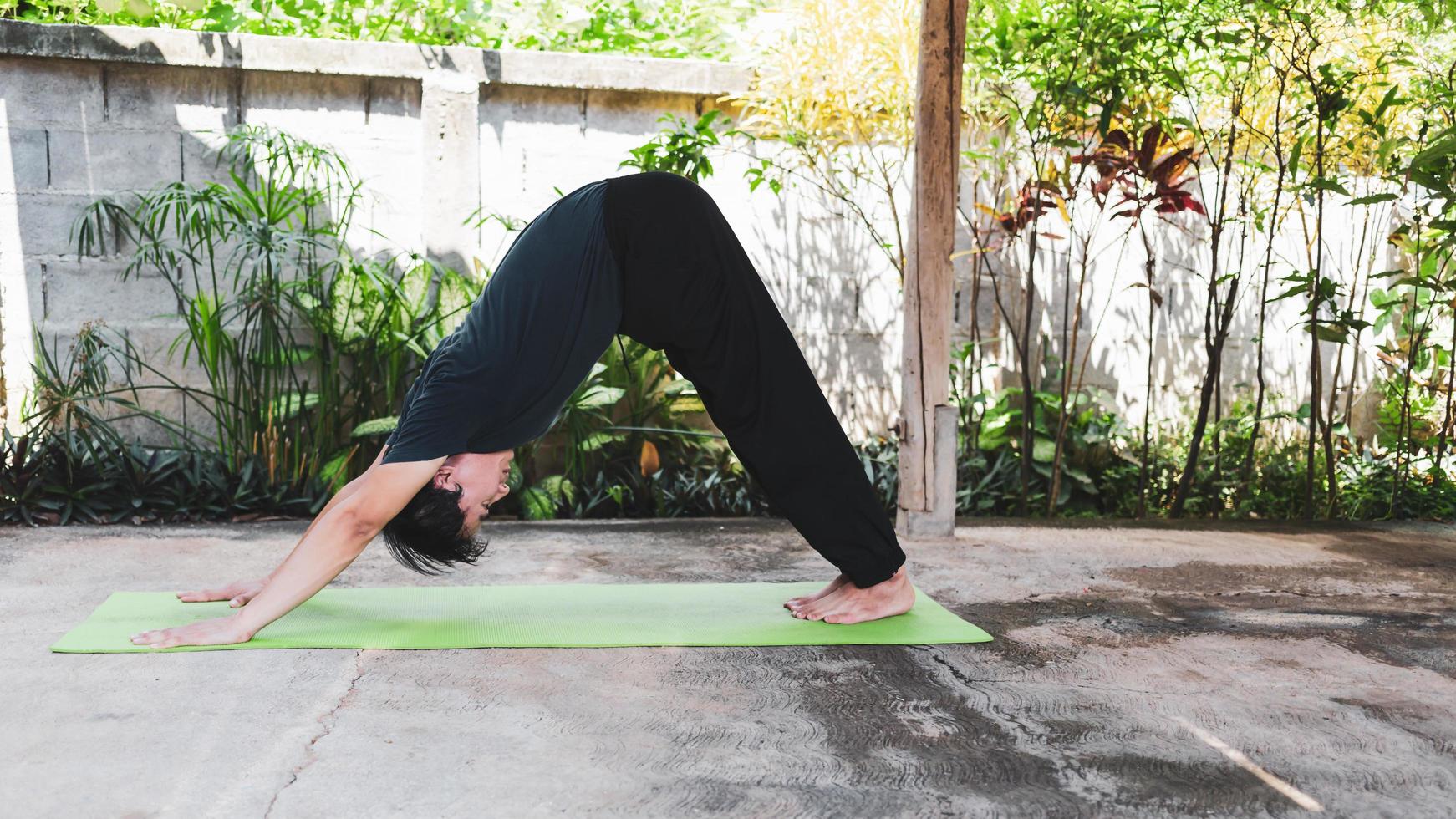 asiatisk man i tillfällig utrusta praktiserande yoga i nedåt vänd hund utgör på de grön yoga matta i de trädgård på Hem. friska livsstil och friska levande begrepp foto