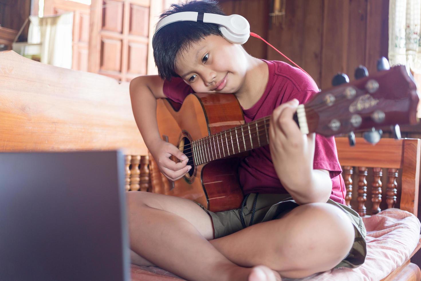 en söt asiatisk pojke bär hörlurar video samtal inlärning musik och skrivning låtar uppkopplad på hans dator bärbar dator använder sig av ett akustisk gitarr lyckligt på Hem unge skapare begrepp foto