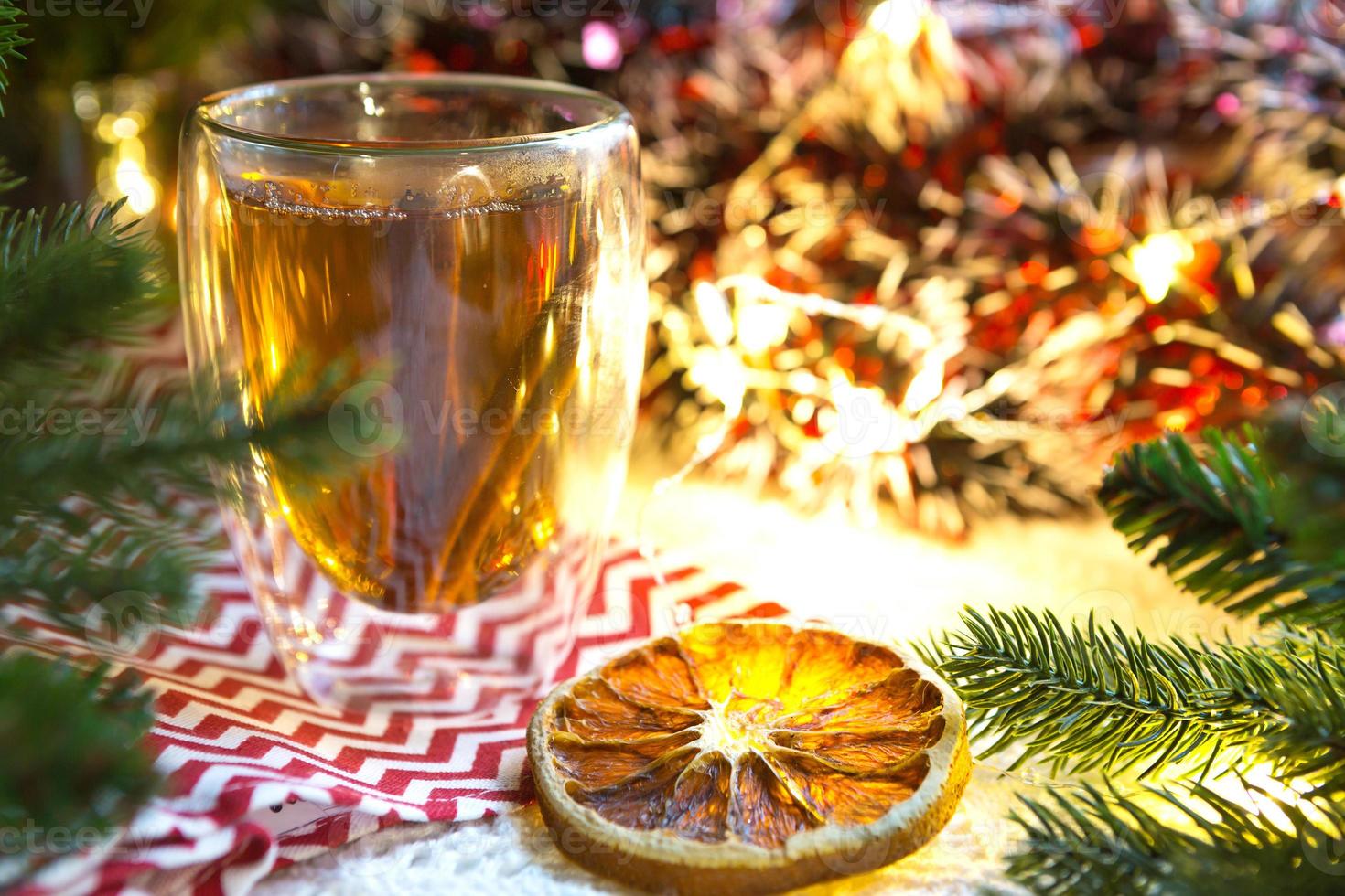 transparent dubbelväggig glas tumlare med varm te och kanel pinnar på de tabell med jul dekor. ny år atmosfär, skiva av torkades orange, krans och glitter, gran gren, mysigt foto