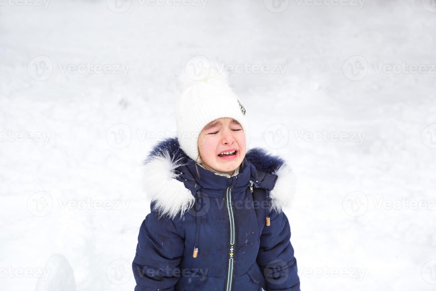 liten flicka gråt utanför i vinter. en barn i värma kläder är upprörd, kall, våtservetter bort tårar, skriker, är nyckfull och hysterisk. vinter, snö, glasera, barns hysteri, missnöje foto