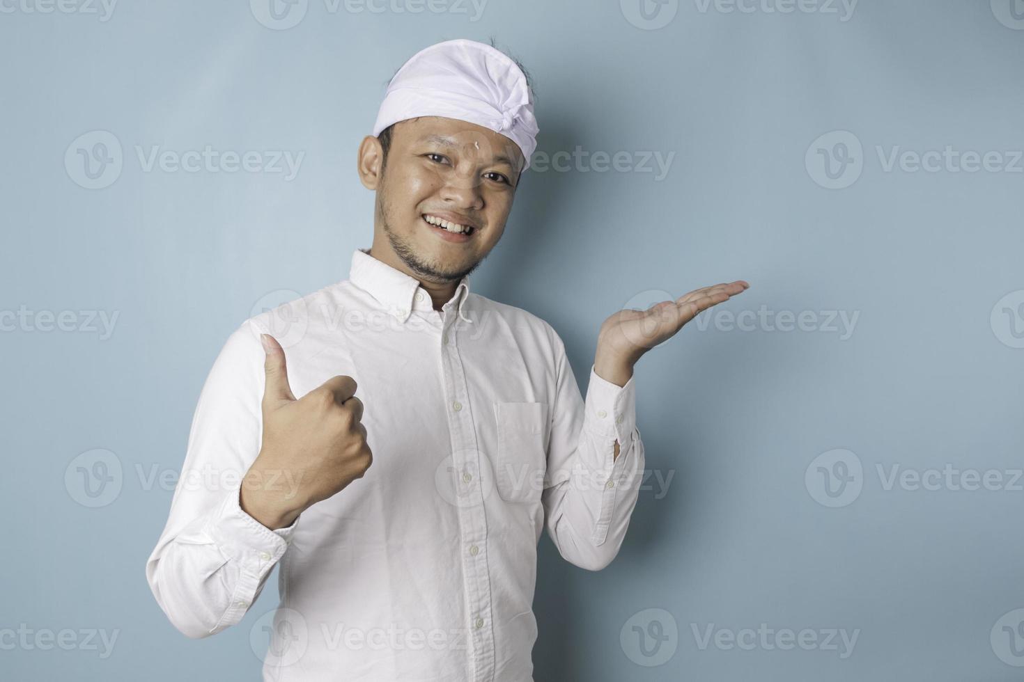 upphetsad balinesisk man bär udeng eller traditionell pannband och vit skjorta ger tummen upp hand gest av godkännande, isolerat förbi blå bakgrund foto