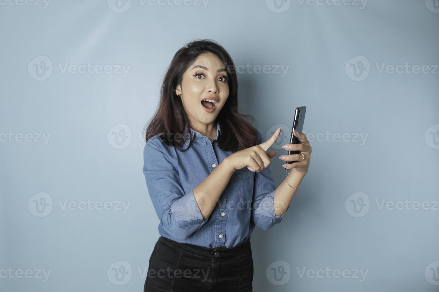 en porträtt av en Lycklig asiatisk kvinna är leende och innehav henne smartphone bär en blå skjorta isolerat förbi en blå bakgrund foto