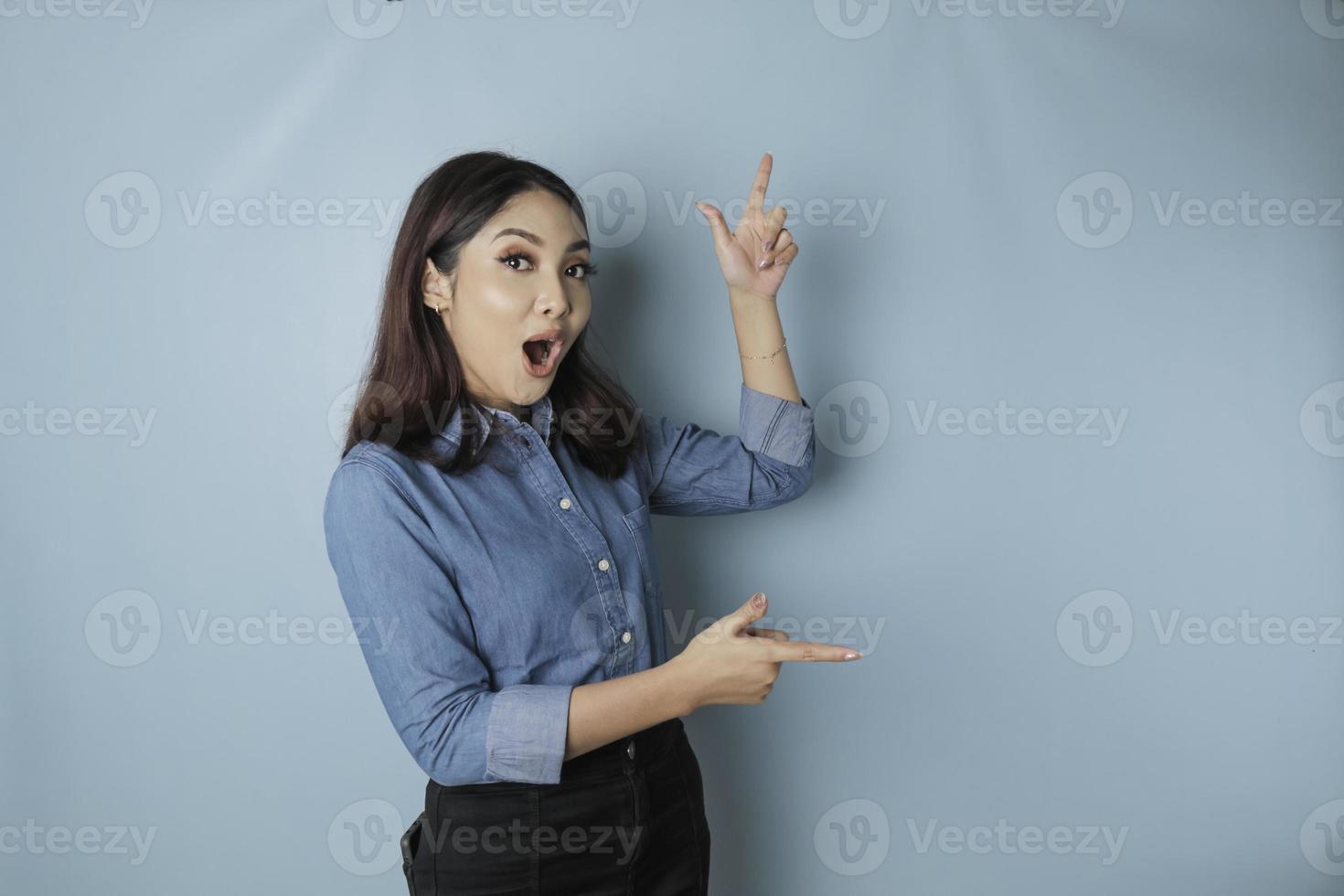 chockade asiatisk kvinna bär blå skjorta pekande på de kopia Plats bredvid henne, isolerat förbi blå bakgrund foto