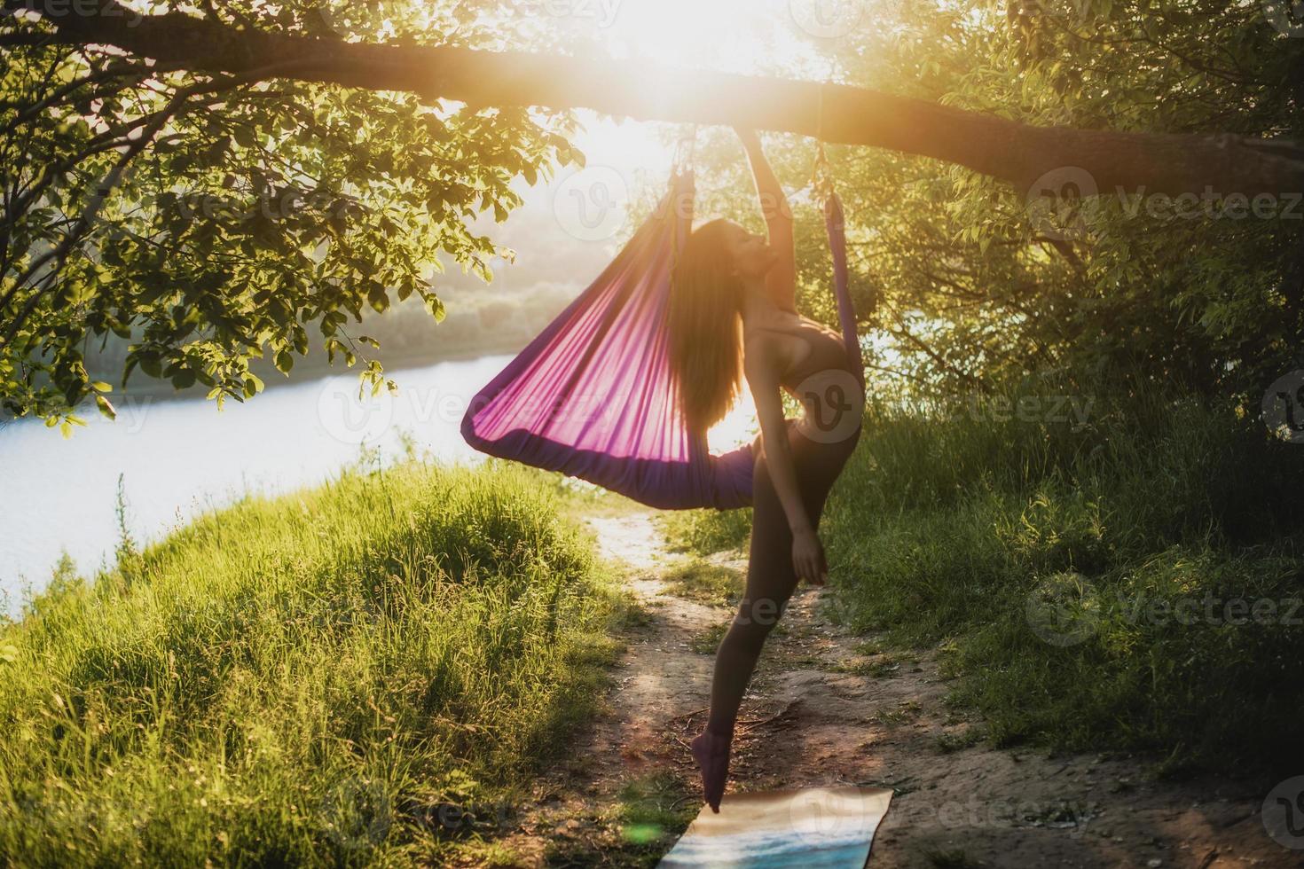 en ung kvinna gymnast är engagerad i antenn yoga, använder sig av en kombination av traditionell yoga poserar, pilates och dansa använder sig av en hängmatta på solnedgång i natur. friska livsstil. foto