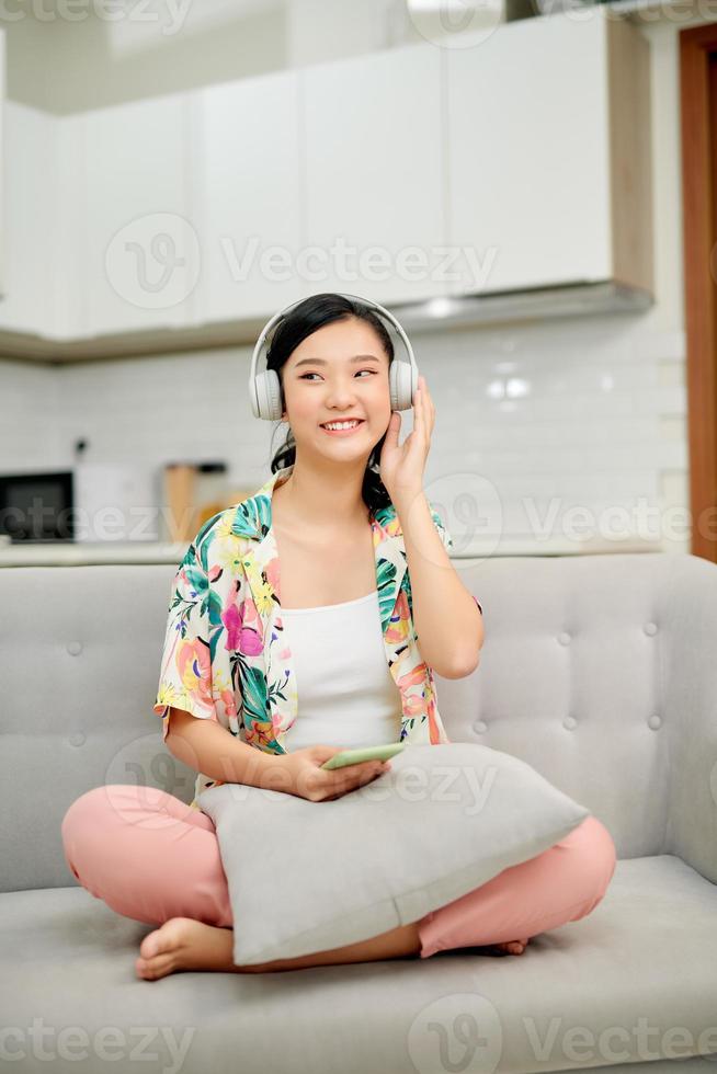 porträtt skön ung asiatisk kvinna använda sig av smart mobil telefon med hörlurar för lyssna musik på soffa i levande rum område foto